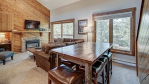 A cozy cabin interior with a wooden dining table, leather seating, a fireplace, and large windows offering a view of the surrounding forest.
