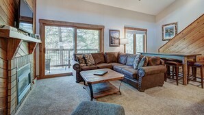 A cozy living room with a sectional sofa, a coffee table, and a fireplace. There are large windows and a wooden staircase.