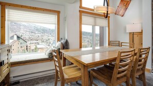 Ein Essbereich mit Holztisch und Stühlen, großen Fenstern mit Jalousien und Blick auf eine Stadt.