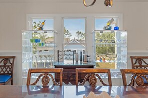 A dining area with a glass table and chairs, a view through large windows, and shelves with glassware and bottles. - Witches Bikini: Private Pool, Hot Tub, Club Access (Corolla)