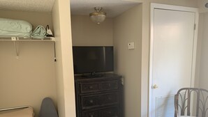 A room with a TV on a dresser, a chair, a door, and a shelf with folded clothes.