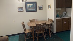 A dining area with a wooden table and chairs, a chandelier, framed pictures on the wall, and a kitchen area with cabinets and appliances.