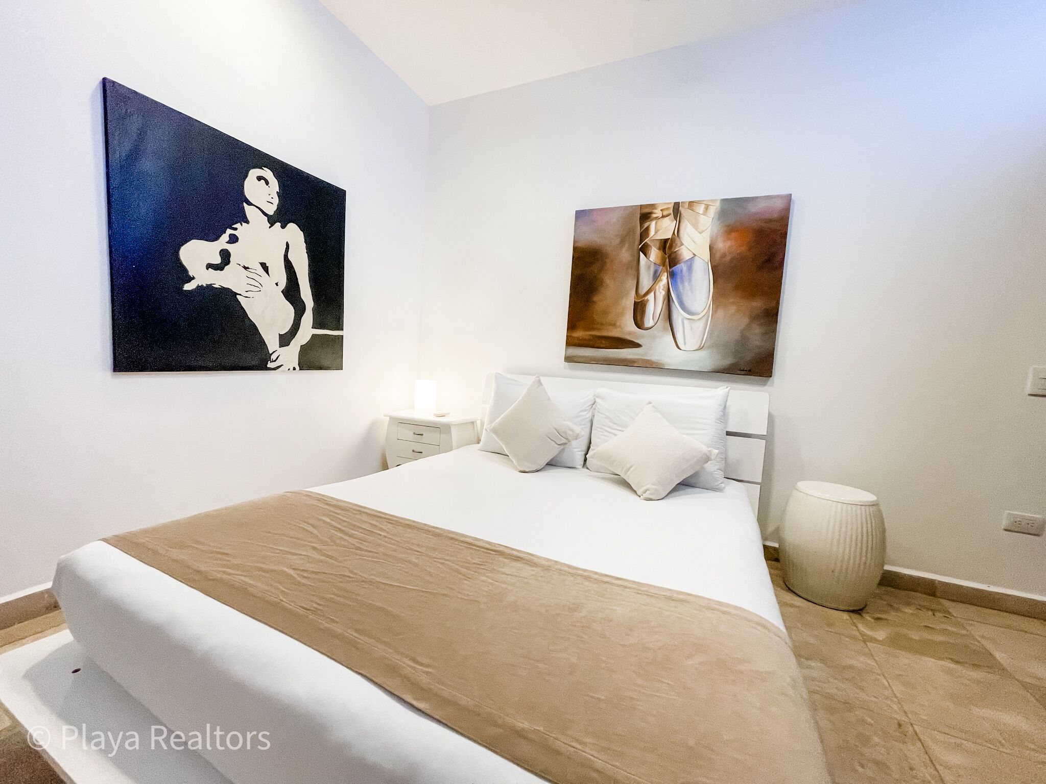 Une chambre à coucher avec un lit, des oreillers blancs, une table de chevet, un vase et deux tableaux accrochés au mur.