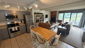 Eine moderne Küche mit Edelstahlgeräten, ein Essbereich mit Holztisch und weißen Stühlen sowie ein Wohnzimmer mit Kamin und großen Fenstern.