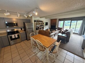 A modern kitchen with stainless steel appliances, a dining area with a wooden table and white chairs, and a living room with a fireplace and large windows. - Snowflake Lodge conveniently located ski in/out sleep 6 Close to Restaurants (Copper)