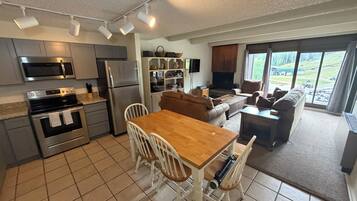 A modern kitchen with stainless steel appliances, a dining area with a wooden table and white chairs, and a living room with a brown leather sofa and a large window offering a view of the outdoors.