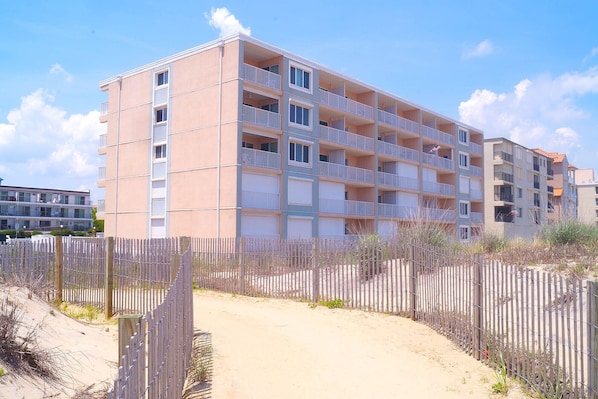 A multi-story building with balconies, a sandy path in front, and a wooden fence.