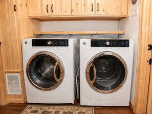 Two front-loading washing machines in a laundry room with wooden cabinets above. - Grizzly Bear - Moonridge | Hot Tub + Fireplace + Forest Views (Big Bear Lake)