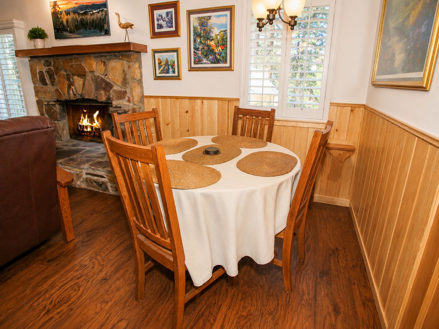 A dining area with a stone fireplace, wooden chairs, a round table with placemats, and framed artwork on the walls.