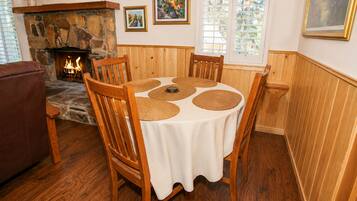 A dining area with a stone fireplace, wooden chairs, a round table with placemats, and framed artwork on the walls.