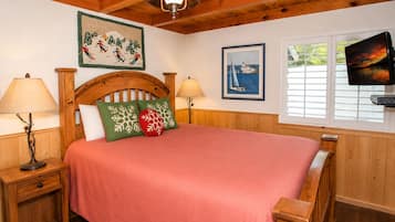 A cozy bedroom with a wooden headboard, a large bed with a red bedspread, two bedside lamps, a framed picture of a sailboat, and a television on the wall.