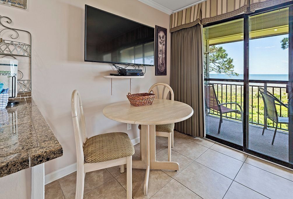 A room with a round table and two chairs, a television mounted on the wall, and a view of the sea through a sliding glass door.
