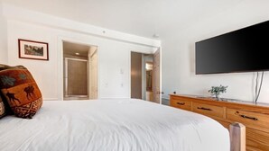 Une chambre à coucher avec un grand lit, une commode en bois, un téléviseur à écran plat et une porte donnant sur une autre pièce.