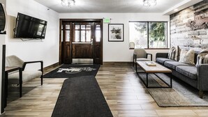 A modern living room with a wooden floor, a sofa, a coffee table, a TV mounted on the wall, and a large window providing natural light.