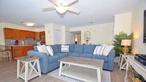 A living room with a blue sectional sofa, a wooden coffee table, and a kitchen in the background.