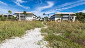 A pathway leading to a beachfront property with modern buildings, surrounded by sand and coastal vegetation. - Sanibel Siesta on the Beach unit 607 (Sanibel)