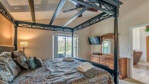 A bedroom with a four-poster bed, a television, a dresser, and a view of the outdoors. - Luxury Pool Home, 2 Master Suites, Lakeview & Games Room. Disney 3 Miles (Kissimmee)