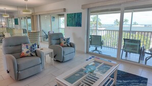 A modern living room with two grey armchairs, a small white table, and a glass coffee table. A large window offers a view of palm trees and a building. - Multiple GULF VIEWs (Sanibel)