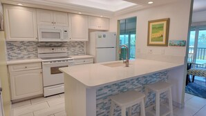 A modern kitchen with white cabinetry, a built-in microwave, stove, and refrigerator. There is a large island with a sink and stools. - Multiple GULF VIEWs (Sanibel)