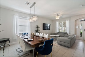 A dining area with a wooden table, chairs, and a chandelier. There is a gray sofa and a small round table in the living area. - Windsor Hills Resort 6 Bedroom Home with Water Views (Kissimmee)