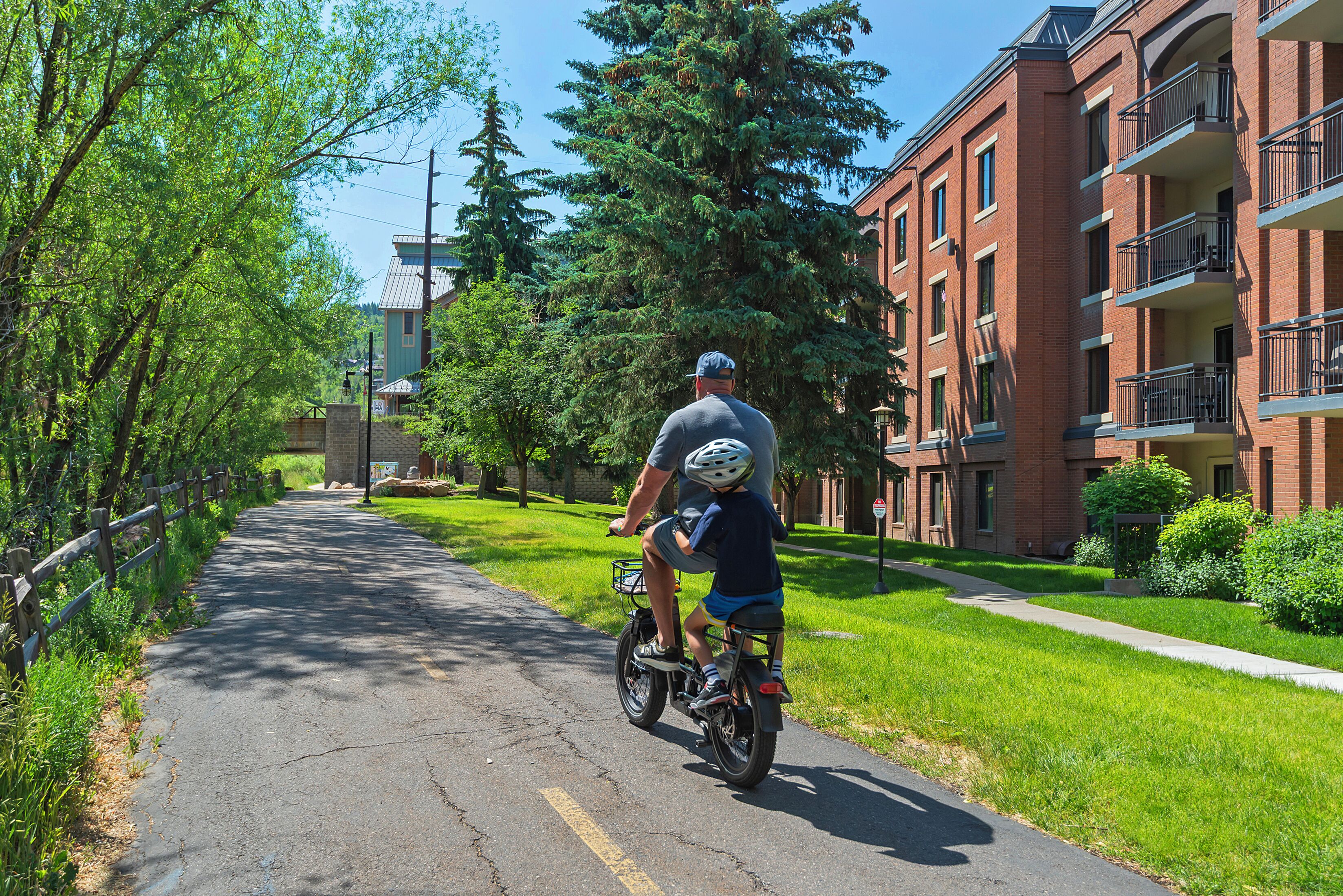Une rue résidentielle où l’on voit un homme et un enfant sur un vélo, des immeubles d’habitation en briques et des arbres verdoyants.