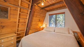Un dormitorio con techo inclinado de madera, una cama con colcha blanca texturizada, una mesita de noche con lámpara y una ventana con vistas a árboles.