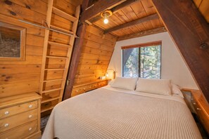 Quarto com teto inclinado de madeira, cama com colcha branca texturizada, mesa de cabeceira com abajur e janela com vista para árvores.