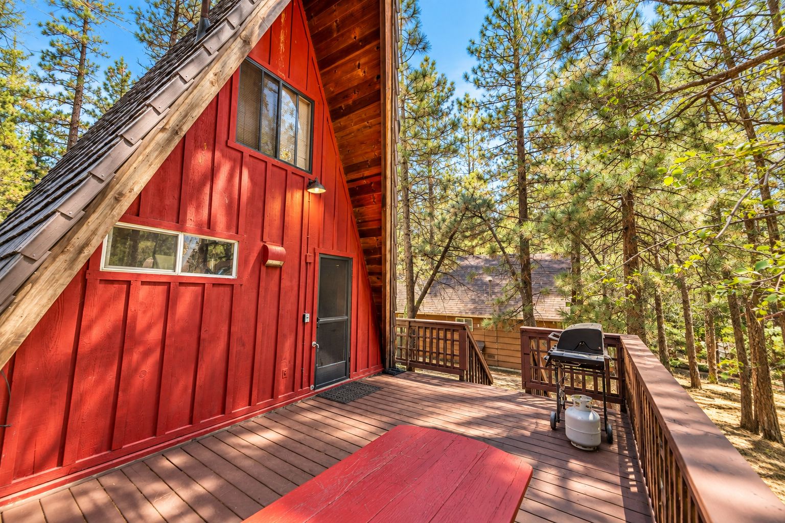 A red cabin with a deck, a grill, and a view of the forest.