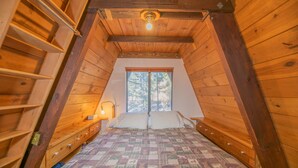 A cozy attic bedroom with wooden walls, a quilted bedspread, and a window with a view of trees. - A-Frame of Mind - Wood Stove Fireplace, Deck, Barbecue (Big Bear City)