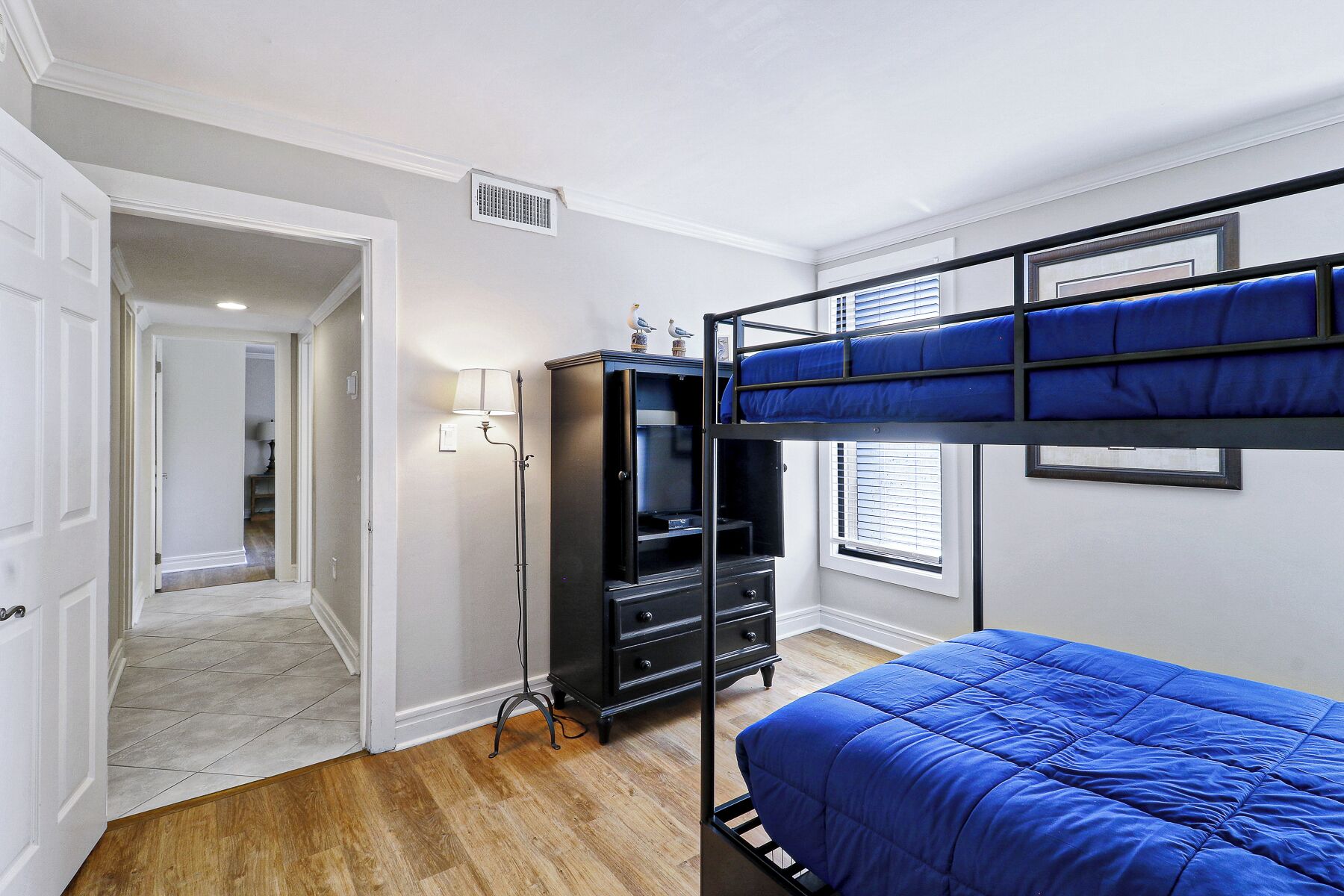 Een stapelbed met een blauwe matras, een zwarte dressoir, een lamp en een raam met jaloezieën.