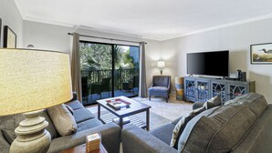 Una sala de estar con sofá, sillón, mesa de centro, televisor y un balcón con mobiliario exterior.