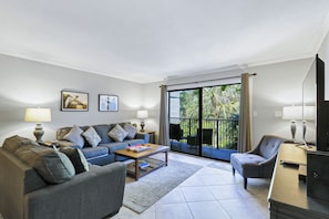 A living room with a sofa, armchairs, a coffee table, and a TV. There are two lamps, a bookshelf, and a sliding glass door leading to an outdoor area with a balcony.