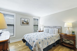 A bedroom with a bed, bedside tables, a lamp, a framed picture, and a window with blinds.