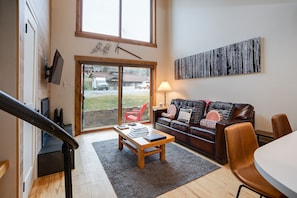 Een woonkamer met een houten salontafel, een leren bank en een groot raam met uitzicht op een gebouw.