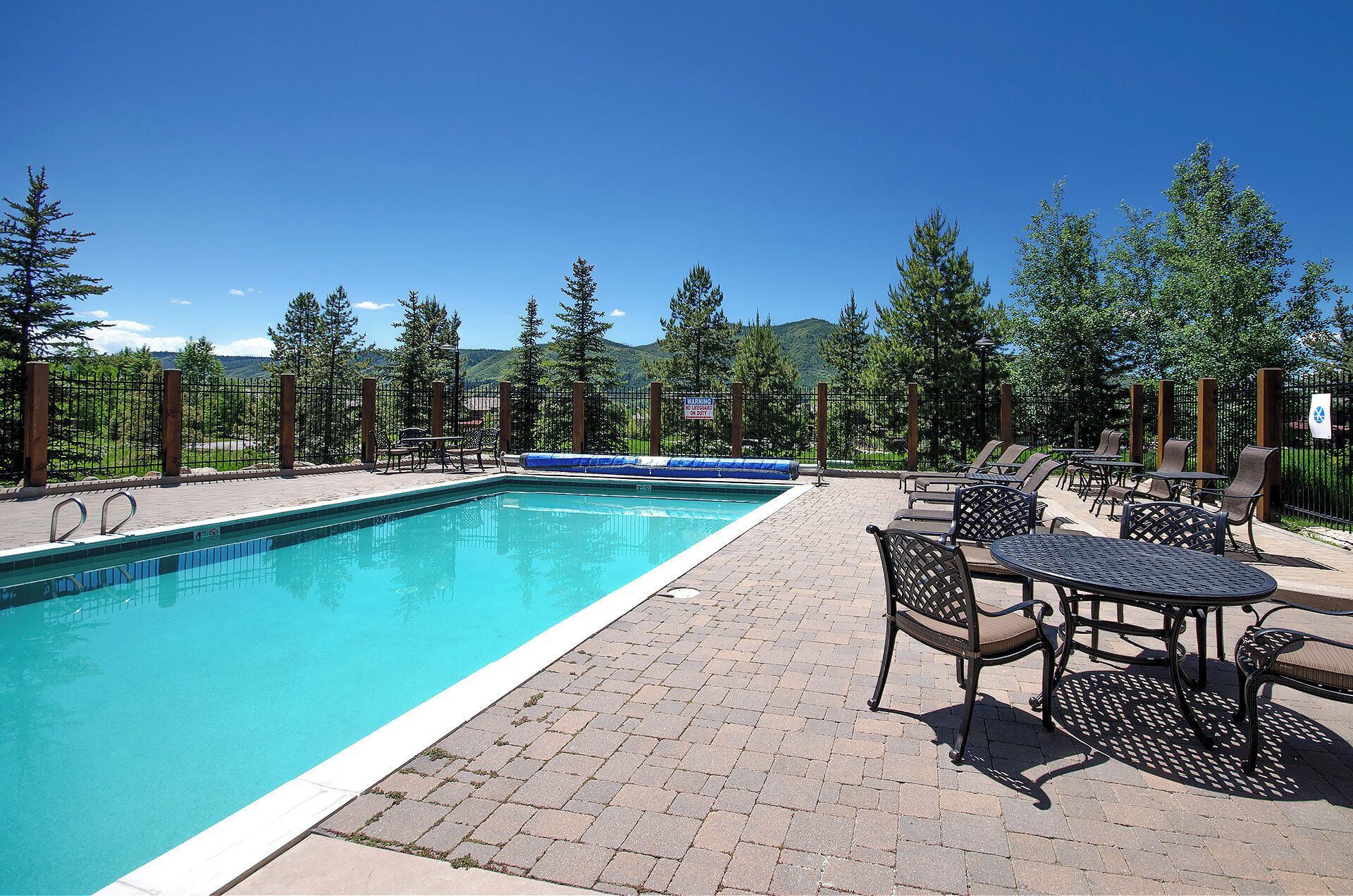 A rectangular swimming pool with a tiled deck, surrounded by outdoor seating and a metal fence.