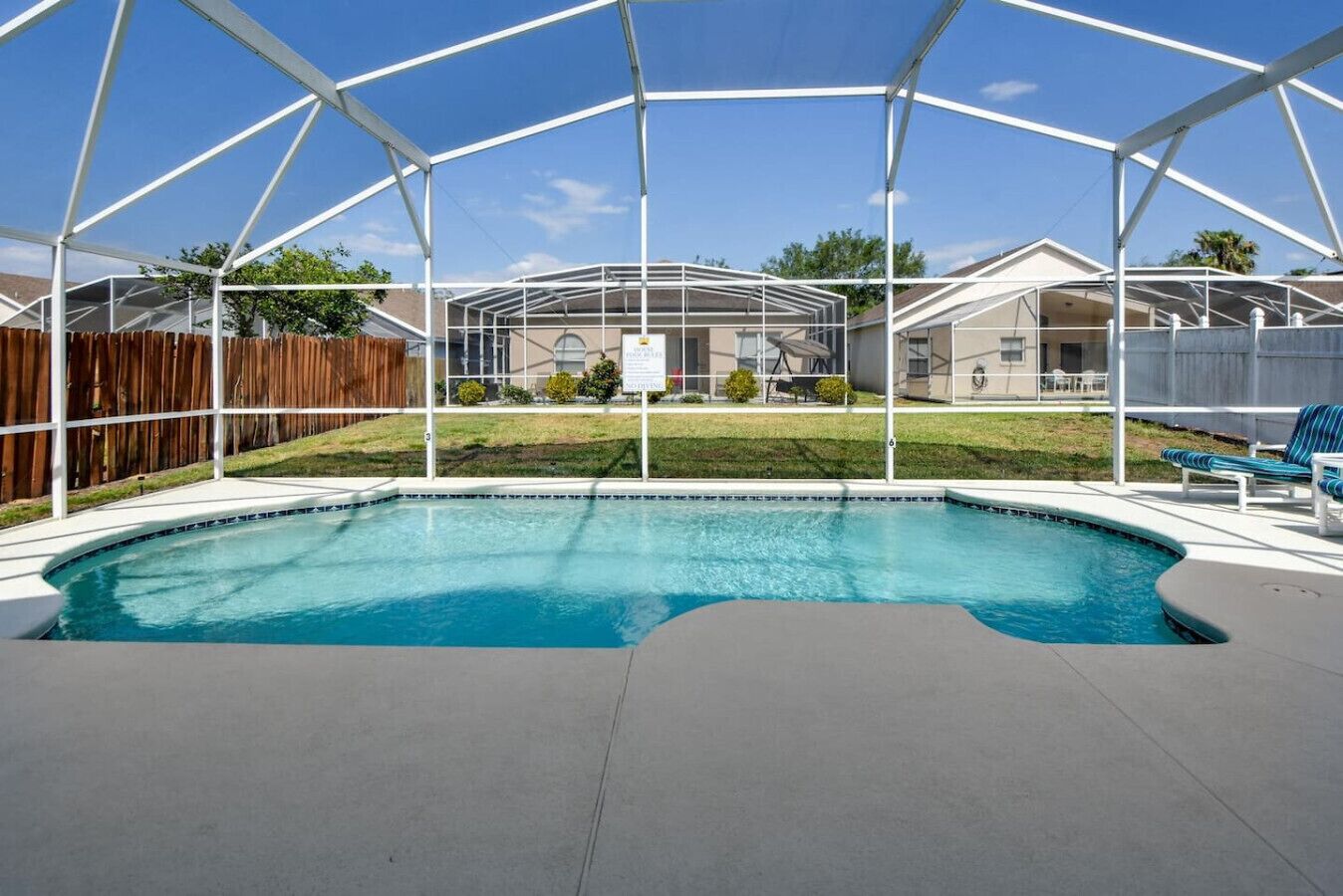 A covered outdoor swimming pool with a clear view of the backyard, including a wooden fence and a house in the background.