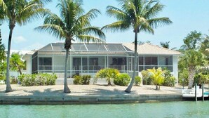 A house with a glass extension, surrounded by palm trees and a body of water. - Now Available! Enjoy Panoramic Water Views, Pool, Dock, Beach Parking Permit & Pup Friendly! (Sanibel)