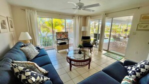 A living room with a sofa, TV, and a balcony with outdoor furniture. - Now Available! Enjoy Panoramic Water Views, Pool, Dock, Beach Parking Permit & Pup Friendly! (Sanibel)