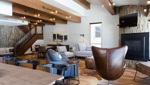 A modern living room with a wooden dining table, leather chairs, a fireplace, and a large TV.