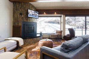 Un salon chaleureux avec une cheminée, des canapés en cuir, un téléviseur à écran plat et une vue sur un paysage enneigé.
