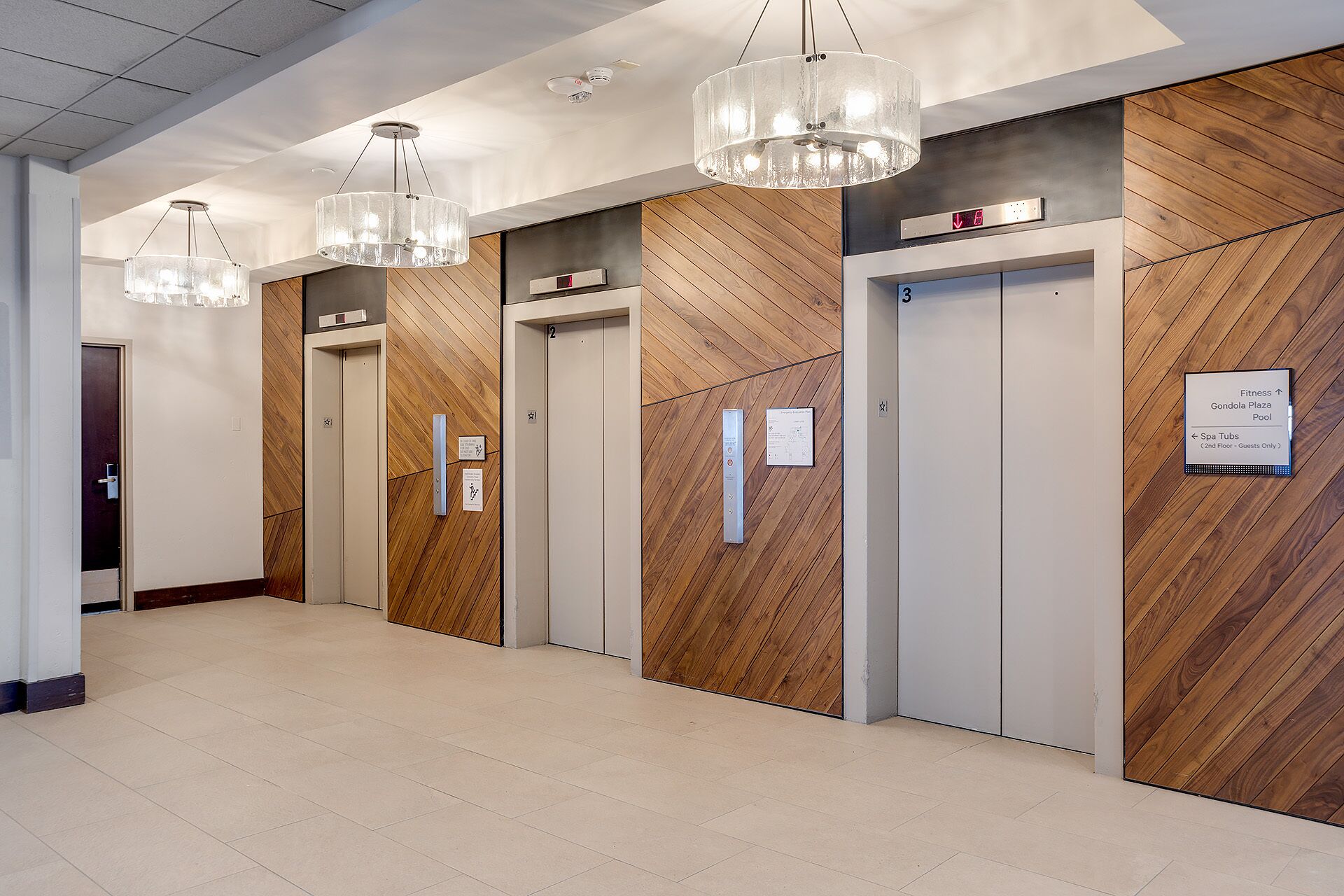 Moderne elevatorområde med træpaneler og tydelig skiltning til 2. og 3. sal, inklusive fitnessområde og spabade.