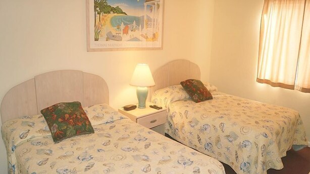 Una habitación con dos camas individuales, una mesita de noche con lámpara, un cuadro en la pared y una ventana con cortinas.