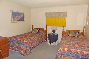 Une chambre avec deux lits simples, une commode en bois, une table de chevet avec une lampe, un tableau encadré au mur et une fenêtre avec des stores jaunes.