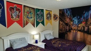 Une chambre avec deux lits, chacun recouvert d’une couette aux couleurs de Poudlard. Quatre bannières représentant les maisons de Poudlard sont accrochées au mur.