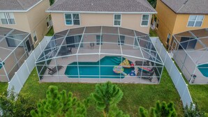 Un quartier résidentiel doté d’une piscine entourée d’une structure en verre, avec des maisons en arrière-plan et de vastes pelouses verdoyantes.