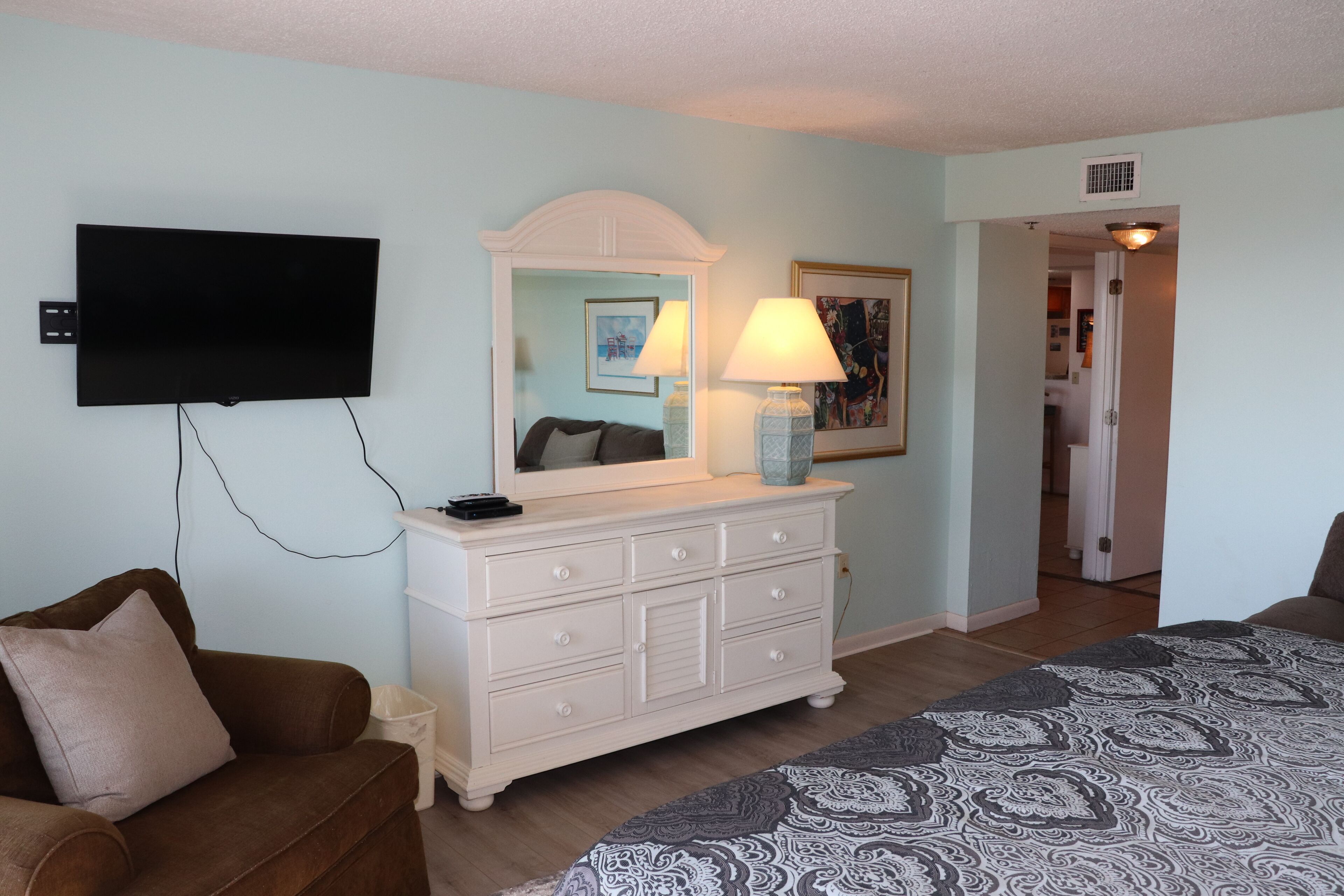 Une chambre à coucher comprenant un lit, une commode, un téléviseur fixé au mur et une porte donnant sur une autre pièce.