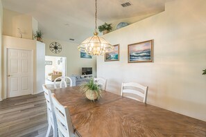 A dining area with a wooden table, white chairs, a hanging light fixture, and two framed paintings on the wall.
