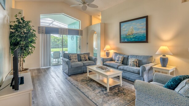 A spacious living room with two sofas, a coffee table, and a TV. There is a large window with a view of greenery outside.