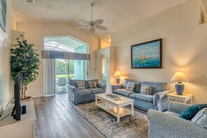 A spacious living room with two sofas, a coffee table, and a TV. There is a large window with a view of greenery outside.