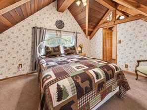 Een slaapkamer met een houten plafond, een bed met een quilt met dessin, een raam met gordijnen en een deur met houten luik.